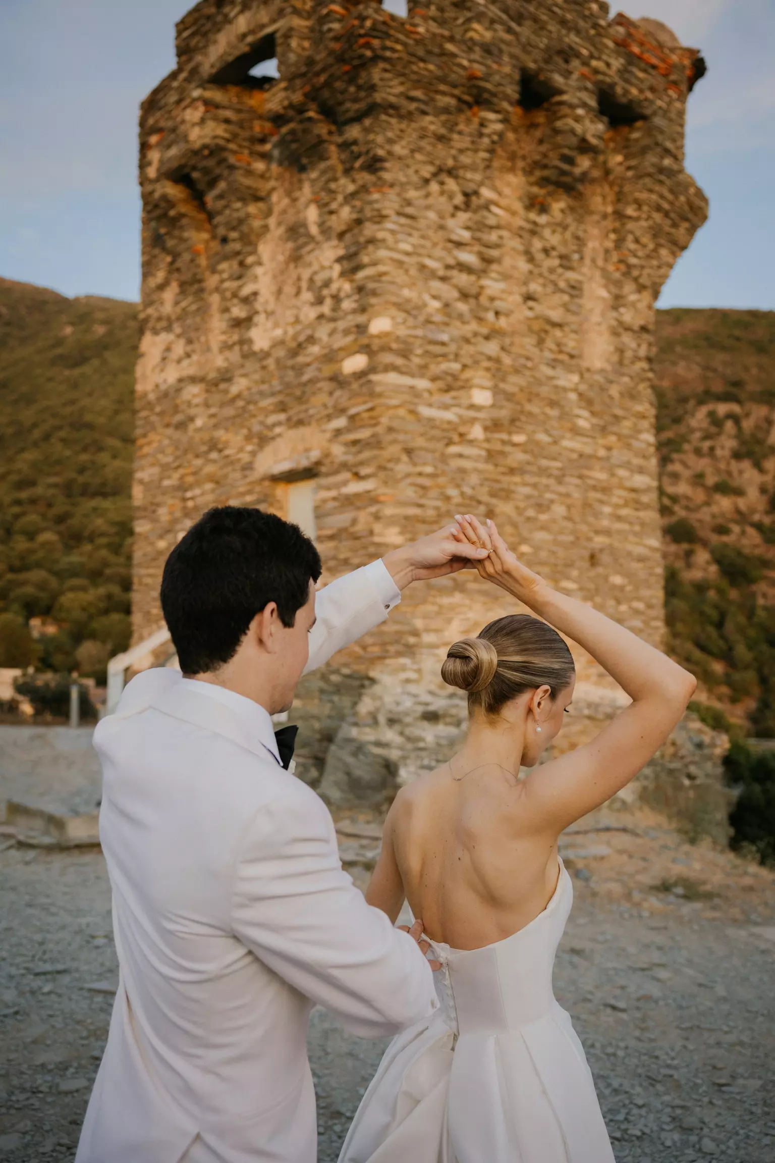 Bride and groom dancing near stone tower in Nonza featuring refined bridal hairstyle by Mandy Dorian