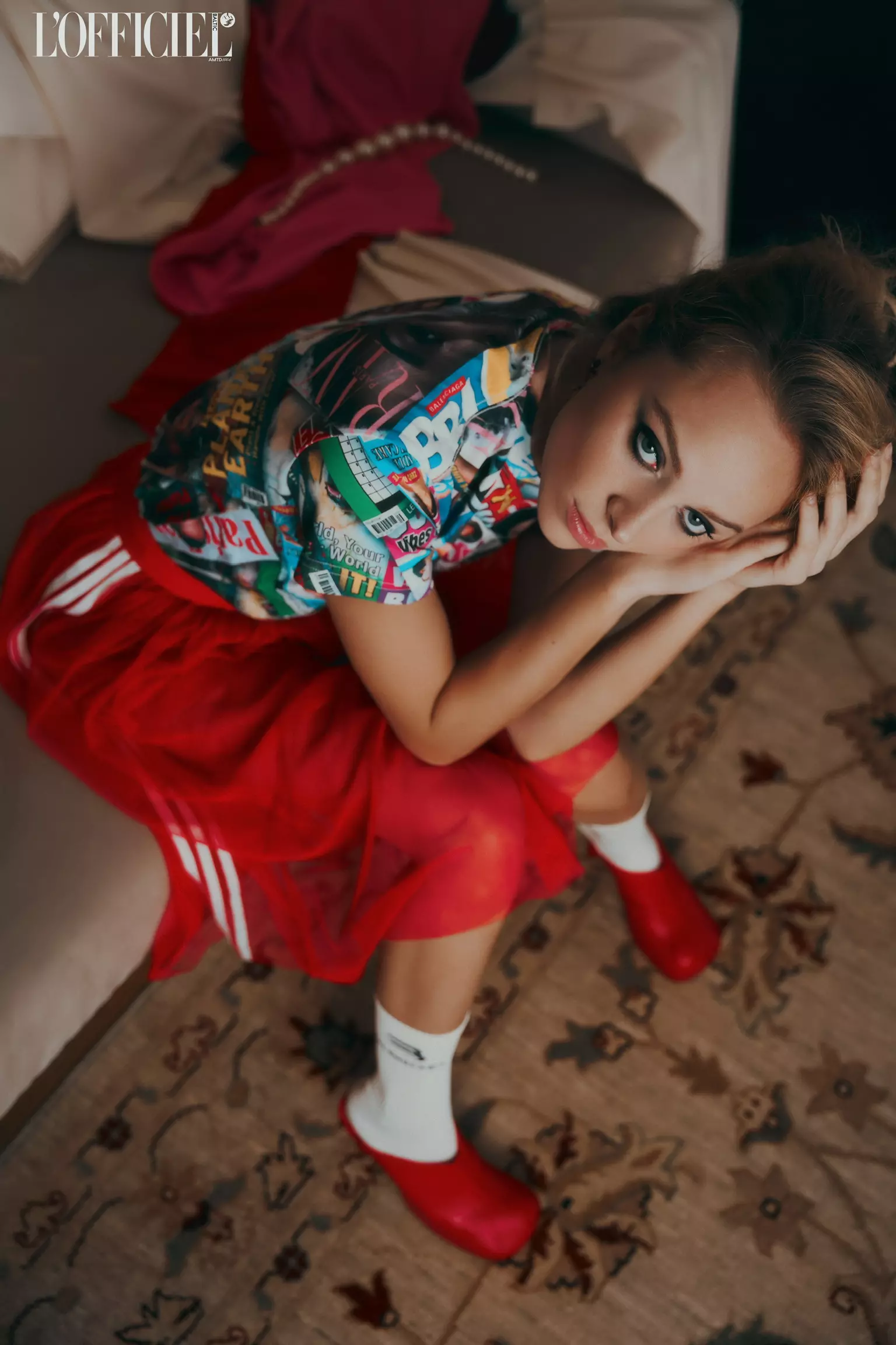 Model seated on patterned floor wearing vibrant outfit with sculpted editorial makeup and styled hair