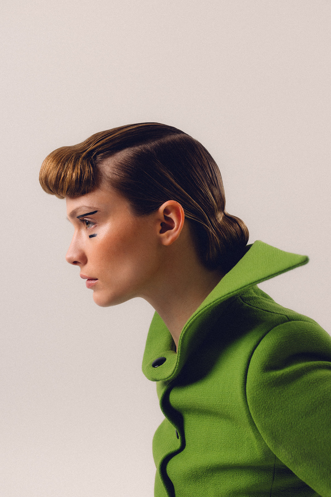 Side profile of model wearing green structured coat with graphic eyeliner and polished hairstyle
